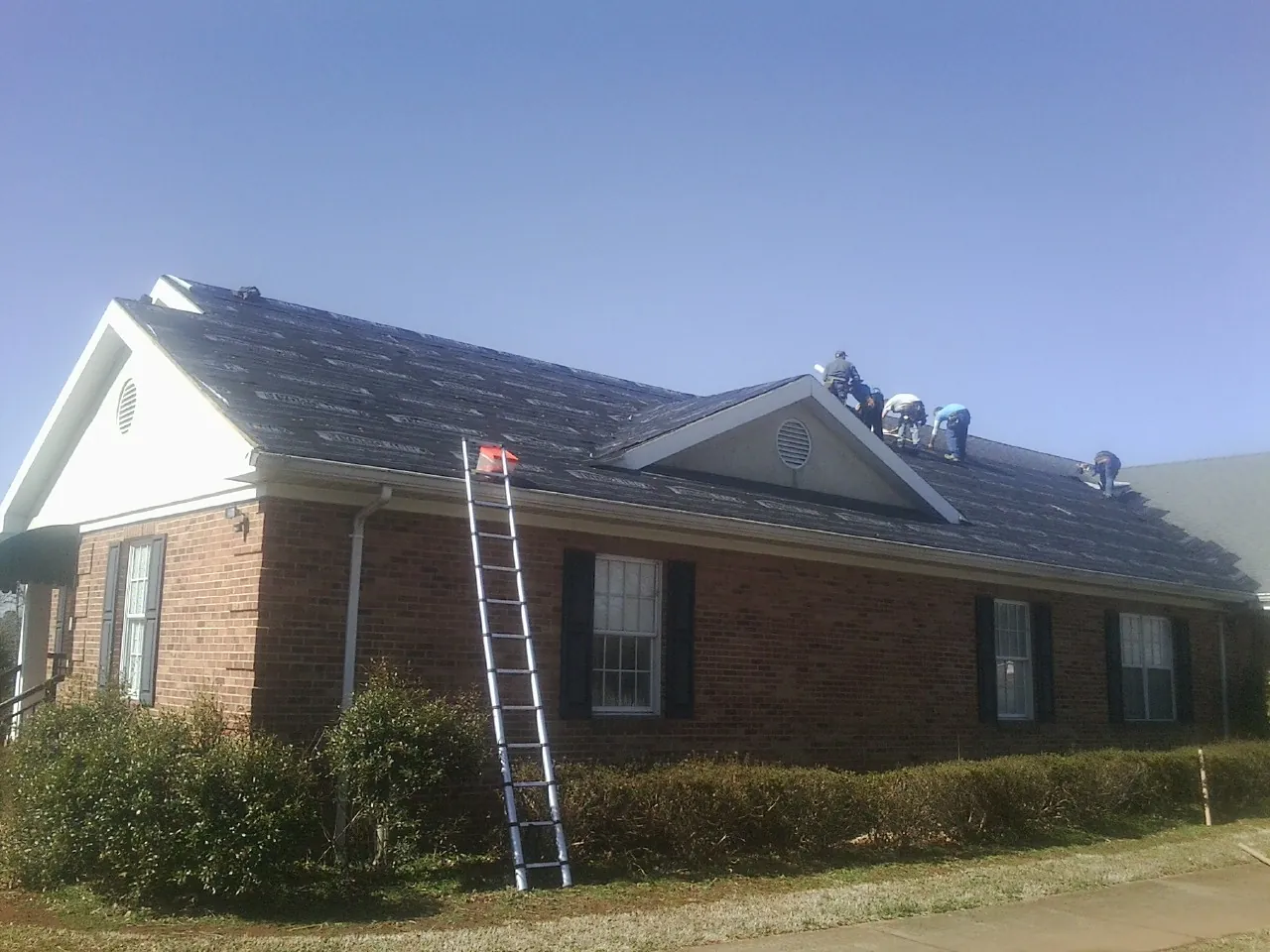 Crew tearing shingles off a brick ranch roof with a ladder set — Metal Roof Repair work by our crew in Oakfield