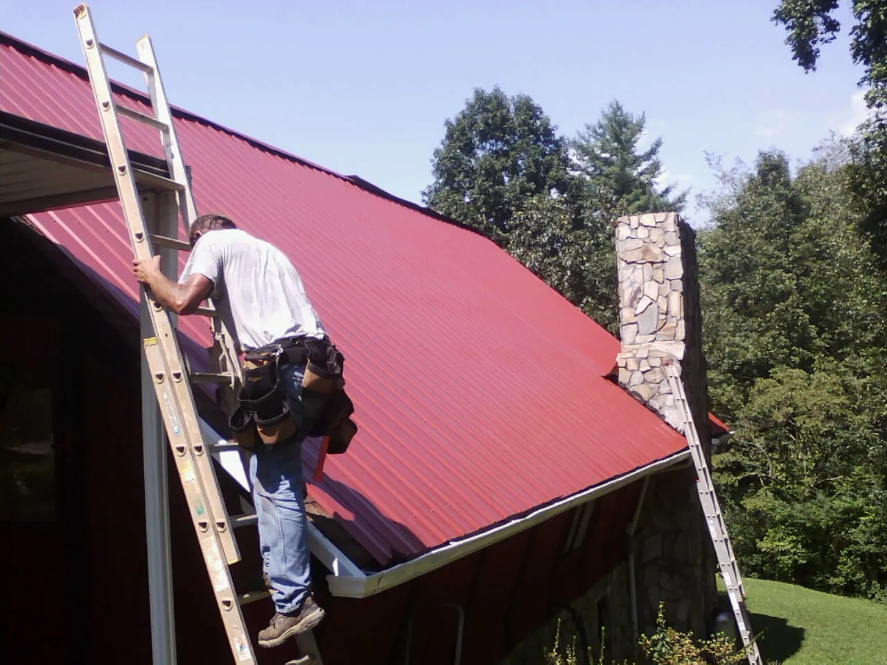 Roofer on a ladder beside a red metal roof and stone chimney — Soffit & Fascia Repair by Roofing Brothers in Oakfield